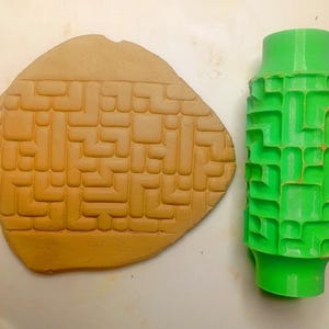 May include: A bright green cylindrical rolling pin with a recessed geometric maze pattern next to a flattened piece of tan clay showing the same embossed maze design, positioned beside a ruler measuring approximately 10 inches in length.
