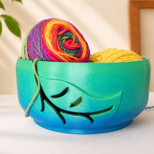 Può includere: Una ciotola per gomitoli rotonda con una sfumatura di colore che passa dal verde in alto al blu alla base. La ciotola presenta un design intagliato a forma di foglia sul davanti, contenente un gomitolo di lana arcobaleno e uno giallo.