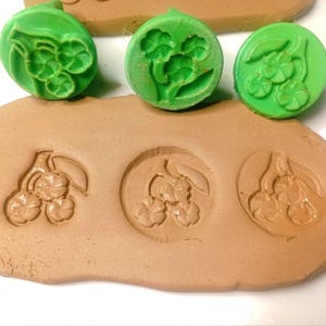 Puede incluir: Tres sellos circulares de plástico verde con un diseño floral en relieve, colocados sobre una pieza plana de arcilla marrón claro que ha sido marcada con los mismos tres patrones florales, creando un conjunto de motivos circulares.