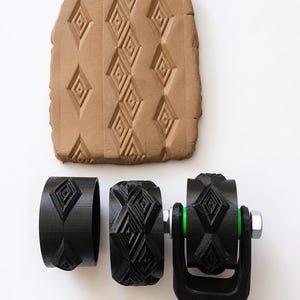 May include: A selection of black 3D-printed clay rollers featuring a diamond pattern, displayed with a clay sample showcasing the design. These rollers are for creating textured surfaces in craft projects.