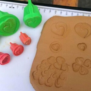 May include: A collection of green and red plastic plunger cutters in various sizes, designed for shaping dough or clay. The tools feature heart and clover motifs, with several impressed patterns visible on a flat, light brown slab of rolled material.