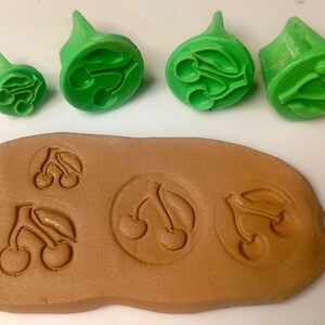 Puede incluir: Cuatro sellos circulares de color verde brillante de varios tamaños, cada uno con un diseño de cereza en relieve. Debajo de los sellos hay una pieza plana y rectangular de arcilla marrón claro con cuatro impresiones de cerezas.