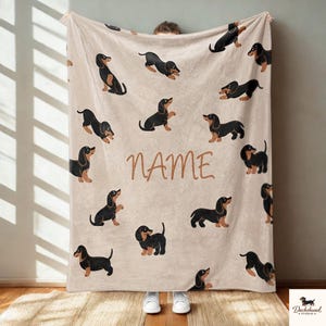 Puede incluir: Una manta beige con múltiples ilustraciones de perros salchicha negros y canela en varias poses. La palabra "NAME" está escrita en escritura marrón en el centro de la manta. La manta está sostenida por una persona que lleva zapatillas blancas.