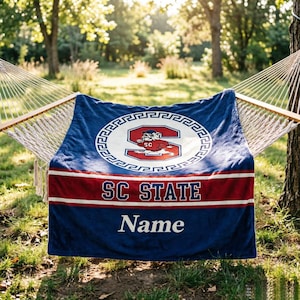 Op de afbeelding: Een blauwe fleecedeken met een wit cirkelvormig embleem met een rode letter S en een bulldog-mascotte. Onder het embleem bevindt zich een rode horizontale streep met de tekst SC STATE in het wit, gevolgd door het woord Name in witte letters.