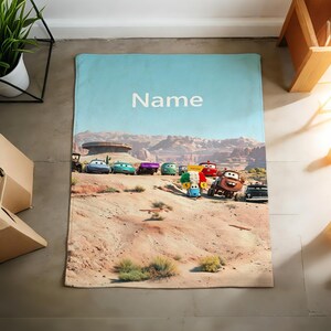 May include: A rectangular blanket featuring a desert landscape with a group of animated car characters from the movie Cars. The sky is light blue with the word Name printed in white text. The scene includes rocky terrain and various colorful vehicles.