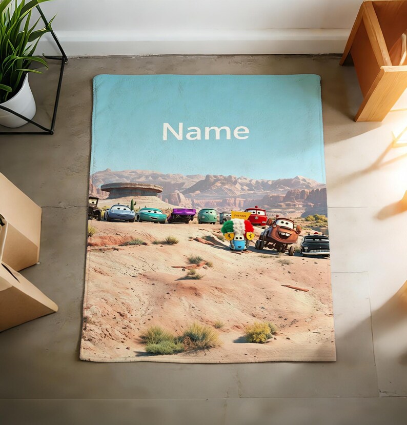 May include: A rectangular blanket featuring a desert landscape with a group of animated car characters from the movie Cars. The sky is light blue with the word Name printed in white text. The scene includes rocky terrain and various colorful vehicles.