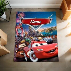 May include: A rectangular blanket featuring a vibrant graphic print of characters from the movie Cars, including a red race car and a brown tow truck. The design includes a large red emblem at the top with the word Name printed in white text.