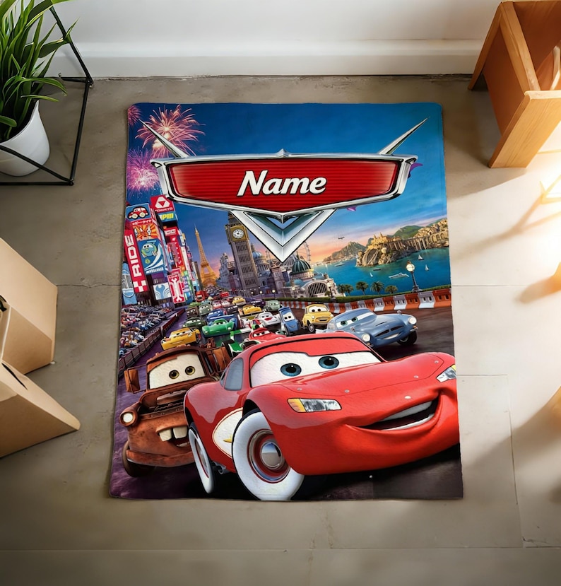 May include: A rectangular blanket featuring a vibrant graphic print of characters from the movie Cars, including a red race car and a brown tow truck. The design includes a large red emblem at the top with the word Name printed in white text.