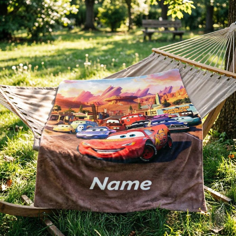 May include: A soft fleece blanket featuring a colorful graphic of Lightning McQueen and other characters from the movie Cars in Radiator Springs. The blanket has a brown bottom section with the white text Name printed in a bold, sans-serif font.