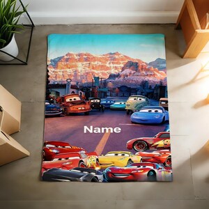 May include: A rectangular fleece blanket featuring a colorful graphic print of various animated car characters from the movie Cars, set against a mountain landscape background with the word Name printed in white text in the center of the road.