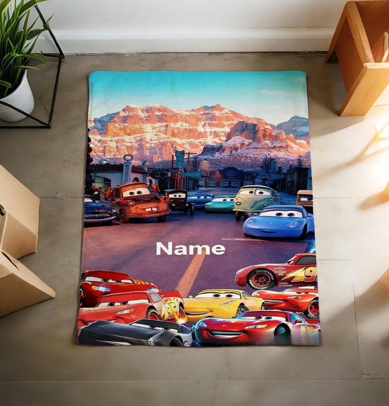 May include: A rectangular fleece blanket featuring a colorful graphic print of various animated car characters from the movie Cars, set against a mountain landscape background with the word Name printed in white text in the center of the road.