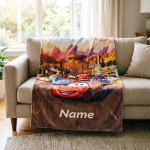 May include: A soft fleece blanket featuring a colorful graphic print of various animated race cars from the movie Cars, set against a desert landscape with the text Radiator Springs and the customizable word Name printed in white at the bottom.