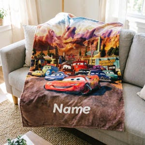 May include: A soft plush throw blanket featuring a colorful graphic print of various animated car characters from the movie Cars, set against a desert landscape background. The word Name is printed in white text at the bottom of the blanket.