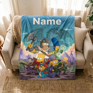 Op de afbeelding: Een lichtblauwe fleece deken met een fantasie cartoon illustratie, inclusief een draak en kasteel. Het woord "Name" is in het wit bovenaan gedrukt. De deken is over een beige bank gedrapeerd.