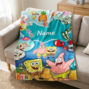 Pode incluir: Um cobertor de lã turquesa com personagens da série animada SpongeBob SquarePants. O cobertor mostra Bob Esponja, Patrick Estrela, Lula Molusco e Sr. Siriguejo, com a palavra "Name" em escrita branca. O cobertor está sobre um sofá cinza.