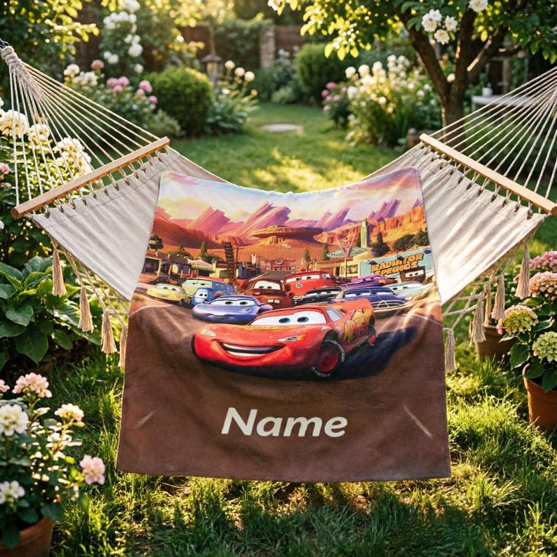 May include: A rectangular fleece blanket featuring a colorful graphic of Lightning McQueen and other race cars from the movie Cars, set against a desert landscape. The word Name is printed in white text at the bottom of the blanket, which is draped over a hammock.