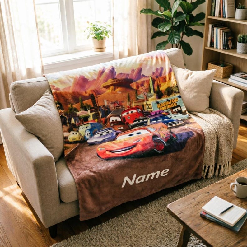 May include: A soft plush throw blanket featuring a colorful graphic print of characters from the movie Cars, including a prominent red race car in the foreground. The blanket has a brown bottom section with the white text Name printed on it.