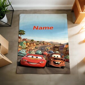 May include: A rectangular fleece blanket featuring a colorful graphic print of the animated characters Lightning McQueen and Mater driving on a road in Radiator Springs. The word Name is printed in bold orange letters at the top of the blanket.