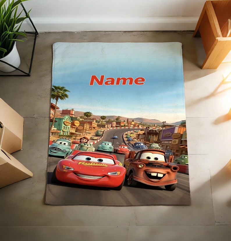 May include: A rectangular fleece blanket featuring a colorful graphic print of the animated characters Lightning McQueen and Mater driving on a road in Radiator Springs. The word Name is printed in bold orange letters at the top of the blanket.