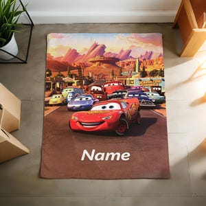 May include: A rectangular blanket featuring a colorful graphic print of Lightning McQueen and other characters from the movie Cars. The scene depicts a desert landscape with mountains and the text Radiator Springs. The word Name is printed in white.