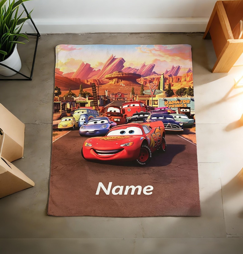 May include: A rectangular blanket featuring a colorful graphic print of Lightning McQueen and other characters from the movie Cars. The scene depicts a desert landscape with mountains and the text Radiator Springs. The word Name is printed in white.