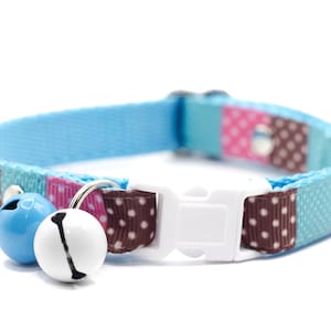 May include: A pet collar featuring a repeating pattern of light blue, pink, and brown segments with white polka dots. The collar includes a white plastic side-release buckle and two small round jingle bells in light blue and white colors.