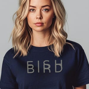 Peut inclure: T-shirt bleu marine avec le mot "BIRD" imprimé en gris clair dans une police stylisée. Le t-shirt est fait d'une matière douce et a un col rond. Le modèle a les cheveux blonds et regarde directement l'appareil photo.