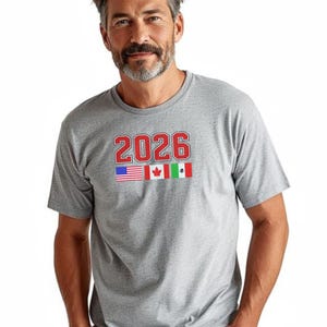 May include: A gray t-shirt with the number "2026" in red block letters. Below the number are the flags of the United States, Canada, and Mexico. The shirt is short-sleeved and has a crew neck.