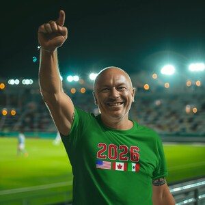 May include: A green t-shirt with the number "2026" in red and white, along with the flags of the United States, Canada, and Mexico. The person is raising their arm in a stadium.