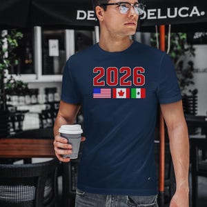 May include: Navy blue t-shirt with the number "2026" in red and white block letters. Below the number are the flags of the United States, Canada, and Mexico. The person in the image is holding a gray coffee cup.