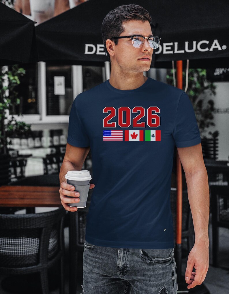 May include: Navy blue t-shirt with the number "2026" in red and white block letters. Below the number are the flags of the United States, Canada, and Mexico. The person in the image is holding a gray coffee cup.