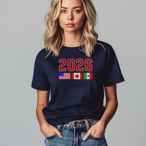 May include: Navy blue t-shirt with the number "2026" in red and white, and the flags of the United States, Canada, and Mexico. The shirt is worn with blue jeans.