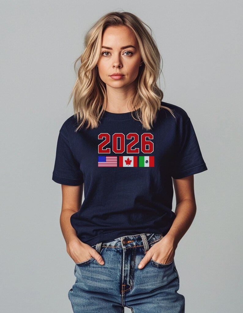 May include: Navy blue t-shirt with the number "2026" in red and white, and the flags of the United States, Canada, and Mexico. The shirt is worn with blue jeans.