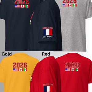 May include: A collection of short-sleeved t-shirts in navy, sport grey, gold, and red. Each shirt features the number "2026" in red, with the flags of the USA, Canada, and Mexico. Some shirts have "WORLD CUP" and "Les Bleus" on the sleeve.