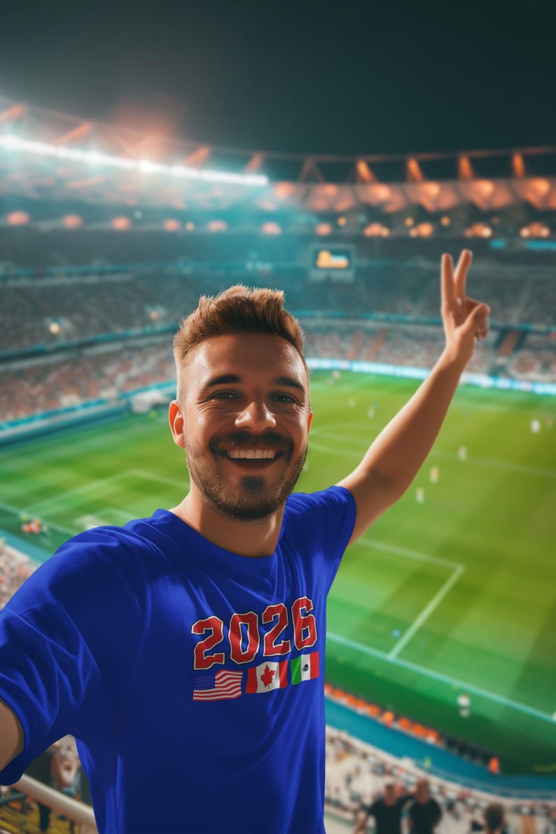 May include: A blue t-shirt with the text "2026" in red and white, along with the flags of the United States, Canada, and Mexico. The person is smiling and giving a peace sign in a stadium.