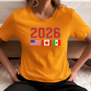 May include: Mustard yellow t-shirt with the number "2026" in red and white block letters. Below the number are the flags of the United States, Canada, and Mexico. The shirt is short-sleeved and made of a soft material.