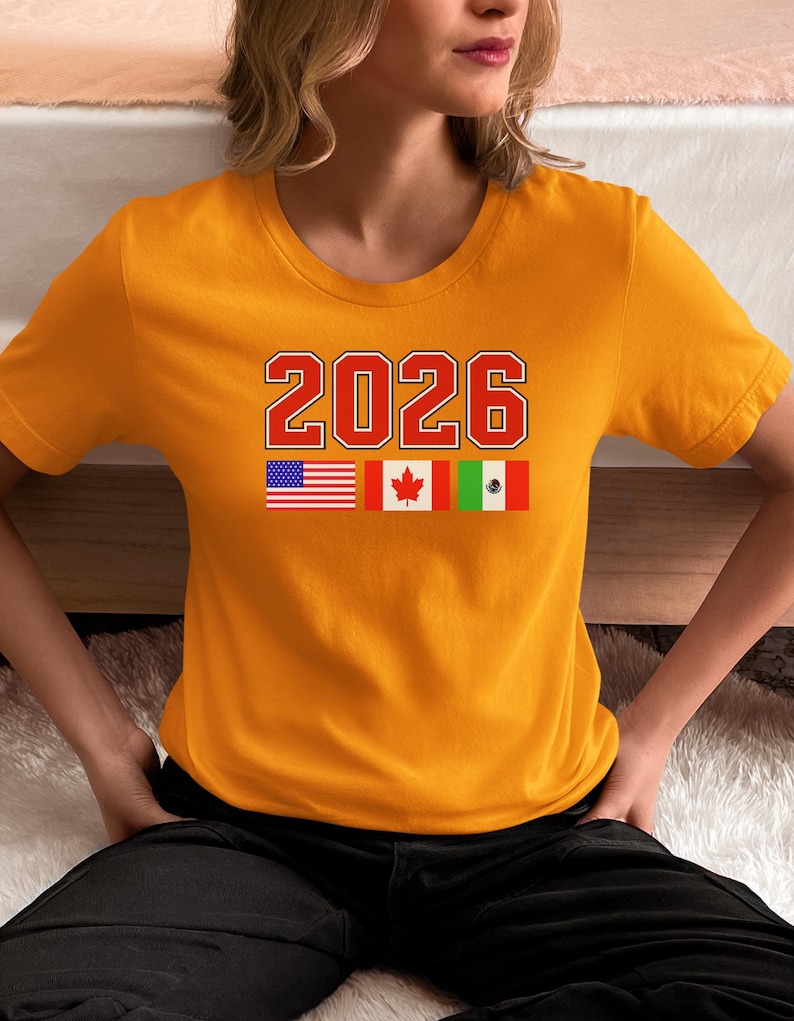 May include: Mustard yellow t-shirt with the number "2026" in red and white block letters. Below the number are the flags of the United States, Canada, and Mexico. The shirt is short-sleeved and made of a soft material.