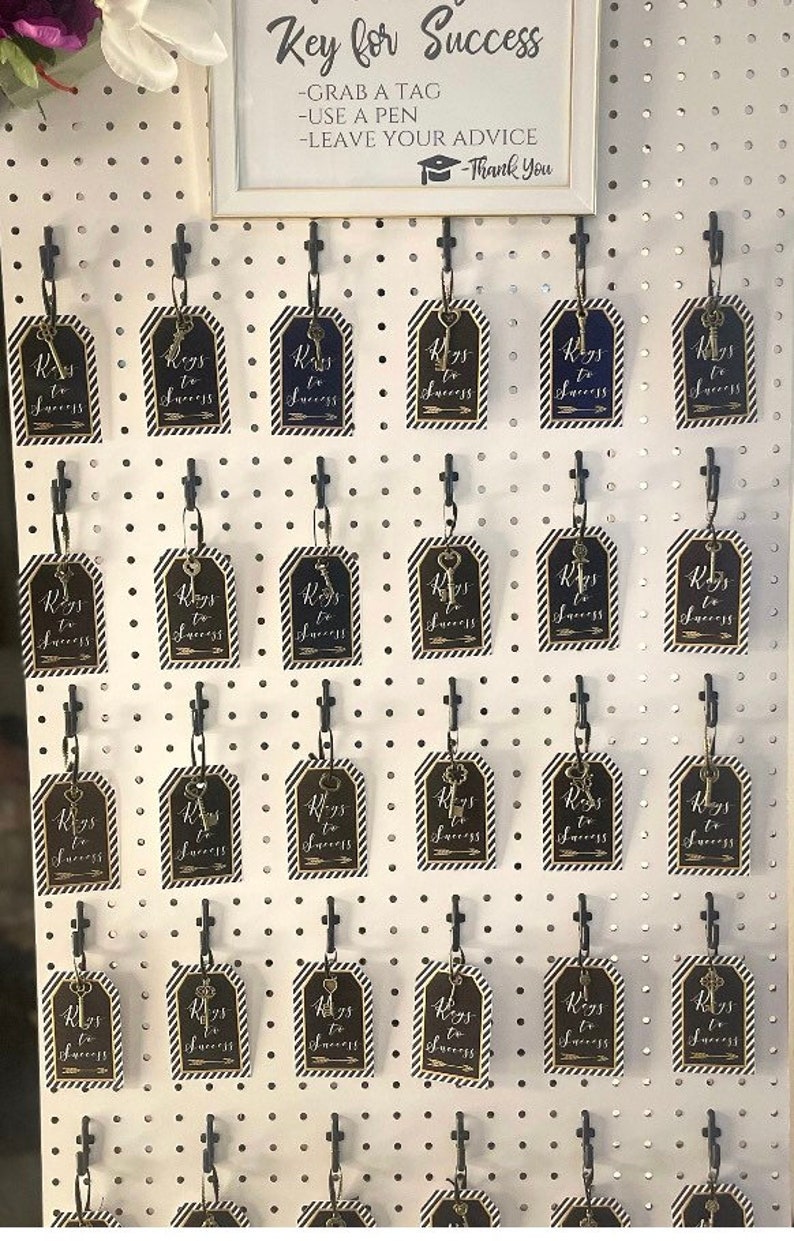 Puede incluir: Un tablero de clavijas blanco con ganchos negros que sostienen etiquetas doradas en forma de llave con el texto "Keys to Success" en cada etiqueta. El tablero de clavijas tiene un letrero encima que dice "Key for Success - Grab a Tag - Use a Pen - Leave Your Advice - Thank You".