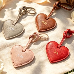 Può includere: Quattro etichette per bagagli in pelle a forma di cuore con cinturini annodati. Le etichette sono grigie, marroni, rosa chiaro e rosse, ognuna con iniziali in rilievo: NR, WH, KM e SA. Le superfici in pelle testurizzata hanno cuciture visibili.