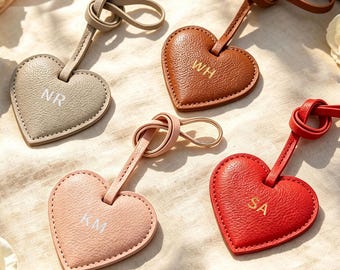 May include: Four heart-shaped leather luggage tags with knotted straps. The tags are colored gray, brown, light pink, and red, each featuring embossed initials: NR, WH, KM, and SA. The textured leather surfaces have visible stitching along the edges.