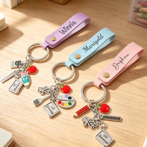May include: Three personalised keychains in lavender, light blue, and light pink, each featuring a metal ring and a leather-like strap with names Winnie, Marigold, and Daphne. Each keychain includes miniature charms of school supplies like pencils, rulers, and apples.