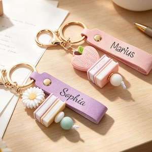 Könnte beinhalten: Zwei Schlüsselanhänger mit goldfarbenen Ringen und Verschlüssen. Einer hat ein rosa Band mit dem Namen Marius, einen rosa Apfelanhänger mit dem Wort TEACH und Bücher. Der andere hat ein violettes Band mit dem Namen Sophia, ein Gänseblümchen und Bücher.