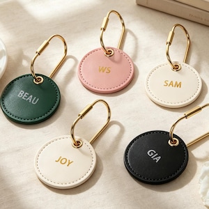 May include: Five circular leather keychains with gold-toned metal loops. The keychains come in dark green, light pink, cream, and black, each featuring gold or silver embossed names or initials: BEAU, WS, SAM, JOY, and GIA, with visible stitching.