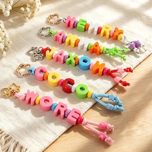 May include: Five colorful beaded keychains featuring the names Henry, Anna, Jaxon, Coco, and Wore. Each keychain is crafted with vibrant plastic letter beads, small round spacer beads, and a gold-toned metal flower-shaped clasp at the top.