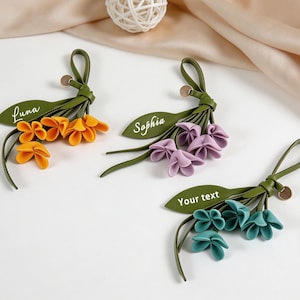 May include: Three floral keychains made of green leather-like material with fabric petals in orange, purple, and teal. Each features a green leaf-shaped tag with white text reading Luna, Sophia, and Your text, plus a small circular metal charm.