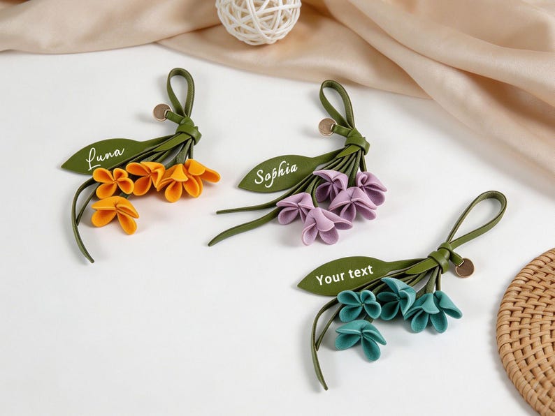 May include: Three floral keychains made of green leather-like material with fabric petals in orange, purple, and teal. Each features a green leaf-shaped tag with white text reading Luna, Sophia, and Your text, plus a small circular metal charm.