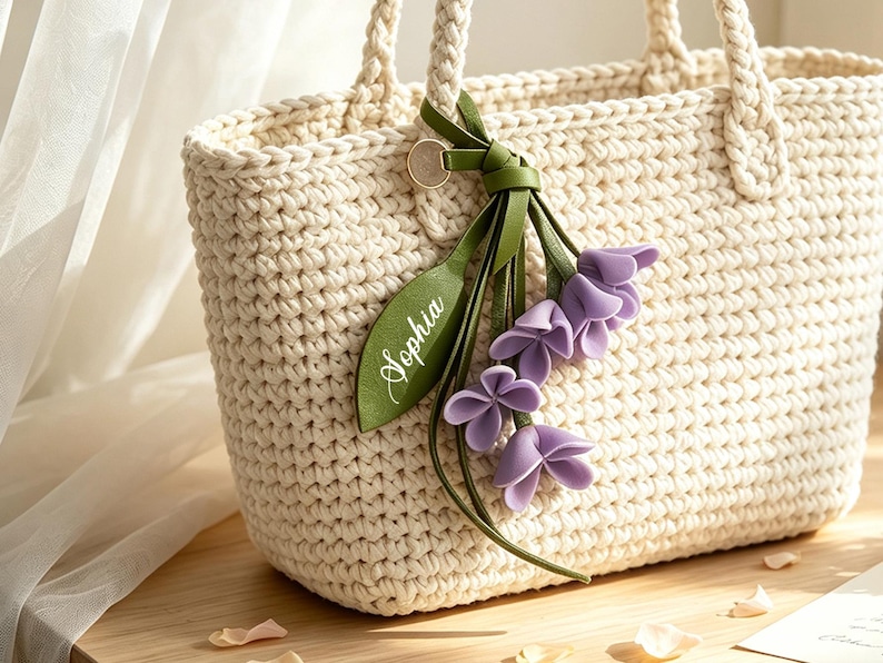 May include: A cream-colored crocheted tote bag with braided handles. Attached to one handle is a green faux leather tag featuring the name Sophia in white script, accompanied by a decorative cluster of three purple fabric flower blossoms.