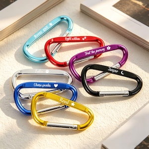 Puede incluir: Una colección de mosquetones de colores en azul, rojo, morado, negro, plateado y dorado. Cada mosquetón tiene una inscripción única, como "Never quit", "Light within" y "Trust the journey". Los mosquetones están dispuestos sobre una superficie clara.