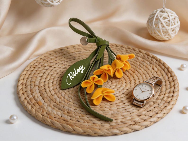 May include: A decorative accessory featuring a green leather-like strap with a loop and a leaf-shaped tag printed with the name Riley in white script. Attached are several small, bright yellow fabric flowers with rounded petals.