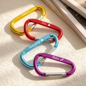 May include: Four colorful carabiner clips in gold, red, blue, and purple. Each clip has a name or phrase engraved in white: "Owen", "Riley", "Keep going", and "Blake". The clips are made of metal and have a spring-loaded gate.